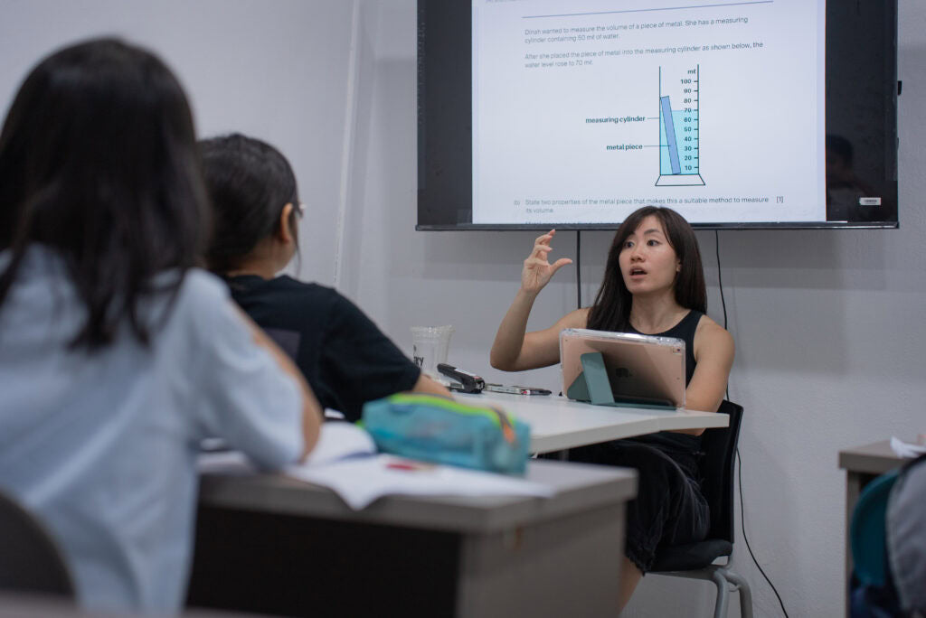A woman in a black sleeveless top sits at a desk in a classroom or office, gesturing toward a large screen that displays a graph. Two people in the foreground are seen from behind, with notebooks and a blue pencil case on the desk.