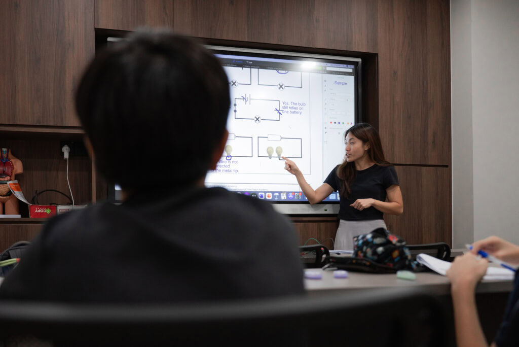 A woman in a black top and white skirt stands in front of a large digital whiteboard showing circuit diagrams, pointing at the screen while a student in the foreground watches from behind.