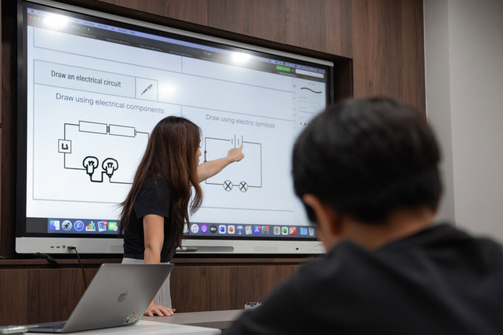 A woman stands in front of a large classroom display screen pointing at an electrical circuit diagram, with a student in the foreground seen from behind and a laptop on the table.